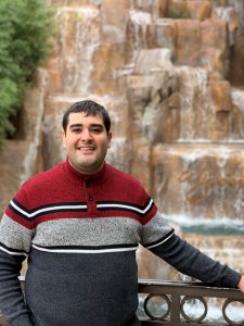 A man smiles at the camera in a red and gray sweater.