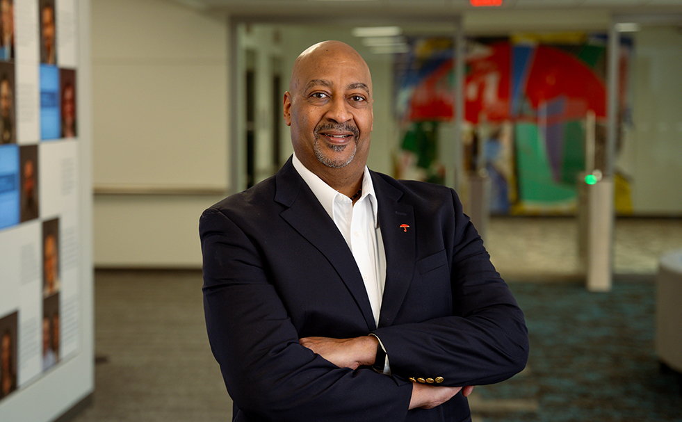 A man in a suit stands in a hallway with his arms folded across his chest as he smiles