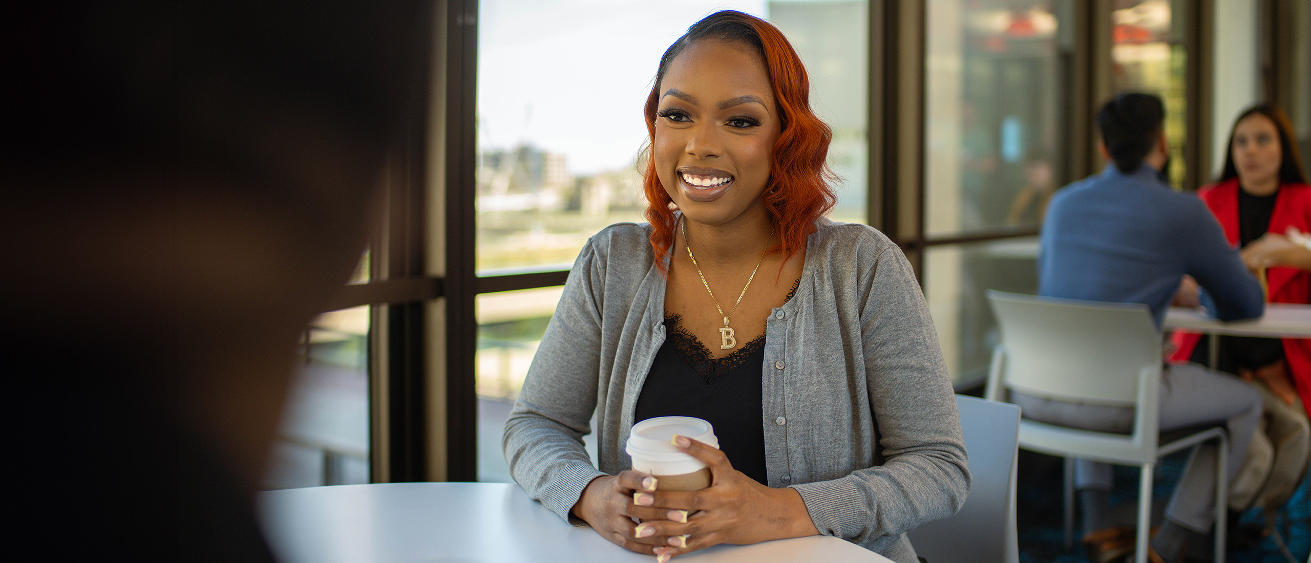 A young woman smiles and sits at a table in front of a window with a coffee cup