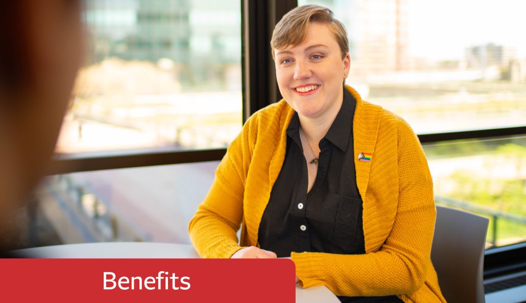 A person in a mustard yellow cardigan with a rainbow pin sits at a table in a bright room with large windows. The background shows a cityscape with buildings and greenery. A red banner with 'Benefits' is in the foreground.