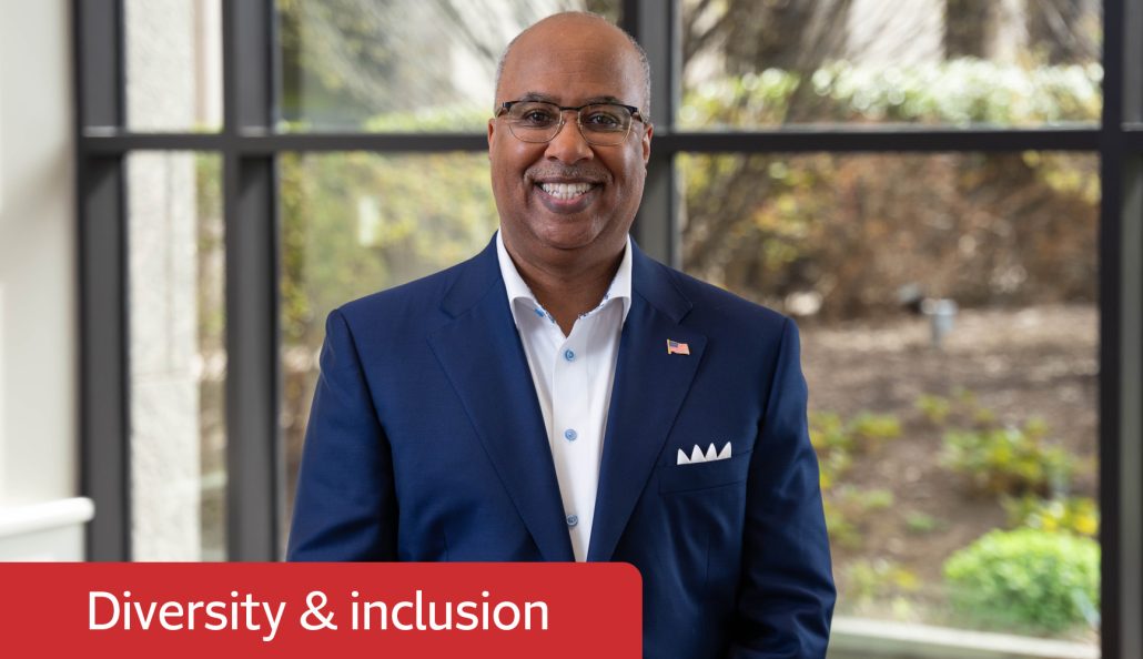 A person in a dark blue suit with a white dress shirt stands indoors near large windows. The suit jacket features an American flag pin and a white pocket square. The background shows a garden with plants and trees. A red banner with 'Diversity & inclusion' is visible in the bottom left corner.