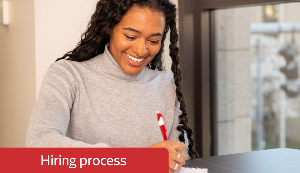 A person with long curly hair sits at a table, writing on a notepad with a red pen. They wear a light gray turtleneck sweater. The indoor setting includes a window with a blurred outdoor view. A red banner with 'Hiring process' is in the bottom left corner.