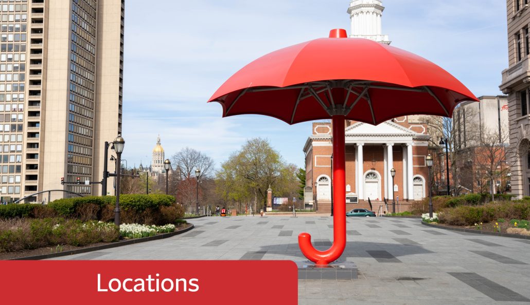 A large red umbrella sculpture is situated in an urban plaza, surrounded by buildings including a modern skyscraper and a historic building with columns. A church with a white steeple and a gold-domed building are in the background. A red banner with 'Locations' is in the bottom left corner.