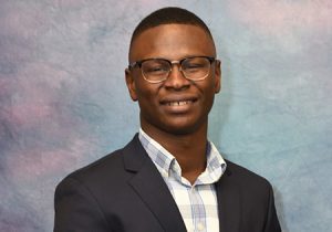 Professional headshot of a smiling man wearing glasses, a dark business suit jacket, and a light-colored checkered or striped dress shirt. The background is a soft, mottled blue and pink gradient, typical of studio photography backdrops. The man has a warm, friendly smile, and the image is well-lit, suggesting it is a formal business portrait.