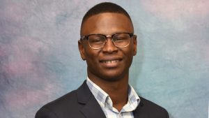 Professional headshot of a smiling man wearing glasses, a dark business suit jacket, and a light-colored checkered or striped dress shirt. The background is a soft, mottled blue and pink gradient, typical of studio photography backdrops. The man has a warm, friendly smile, and the image is well-lit, suggesting it is a formal business portrait.