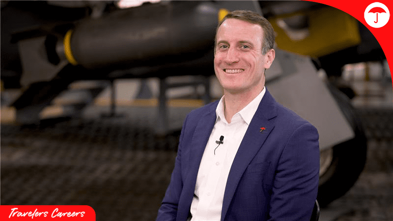 Professional headshot of Caleb Earnest, Chief Underwriting Officer at Travelers, smiling confidently in a navy blue blazer and white collared shirt. He is photographed in an aviation facility with aircraft equipment visible in the blurred background. The image features Travelers Careers branding in the bottom left corner and the Travelers logo in the top right corner.