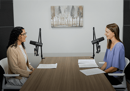 Two women sitting across from each other at a wooden table in a podcast recording studio, each with a microphone on a boom arm. Papers are on the table between them. A landscape painting hangs on the white wall behind them.