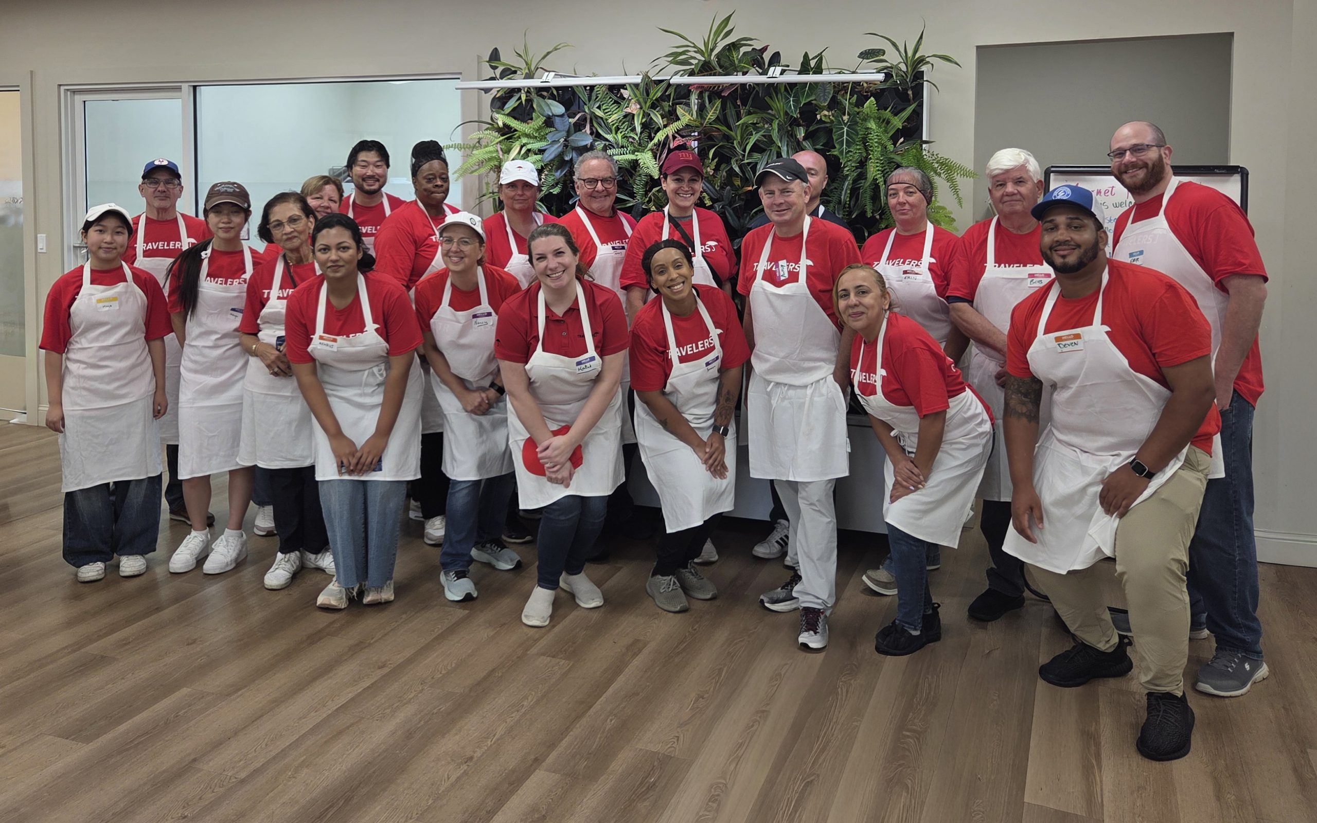 A diverse group of approximately 20-25 volunteers is gathered in a community center or institutional kitchen space. They are wearing matching red shirts and white aprons, indicating participation in a community service activity, likely related to food preparation or serving. The group is arranged in two rows, with some standing and others kneeling or sitting in front. The setting features modern decor with light wood flooring, white walls, and large windows or glass doors. Potted plants are visible along the back wall. The volunteers are smiling, creating a friendly and positive atmosphere, suggesting a regular volunteer activity or special community service event.