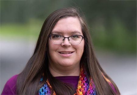 A woman with long, straight brown hair and glasses is smiling warmly. She is wearing a purple top and a vibrant, multicolored crocheted or knitted scarf with bright hues of blue, purple, orange, and yellow. The background is softly blurred, suggesting she is outdoors with greenery or trees behind her. The lighting is natural, giving the photo a professional quality.