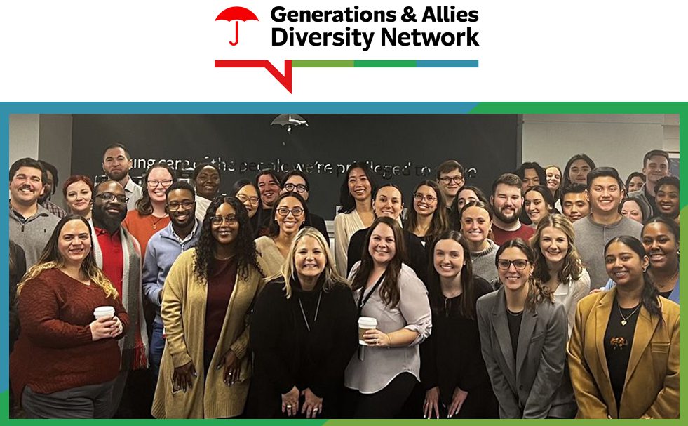 Generations & Allies Diversity Network Group photo of diverse professionals at a corporate event, associated with the Generations & Allies Diversity Network. Approximately 30-40 people of various ages, ethnicities, and backgrounds are dressed in business casual attire, smiling and engaged. The logo for the network, featuring a red umbrella icon and colorful horizontal bar, is displayed at the top. The setting is an office environment with a dark wall in the background.