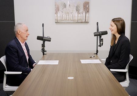 Two people sitting across from each other at a wooden conference table in a professional interview or podcast recording setup. On the left is an older man with gray hair wearing a dark suit, and on the right is a younger woman with long brown hair wearing a black blazer. Both have microphones positioned in front of them and papers on the table. The room has white walls with framed artwork and appears to be a modern office or studio space.