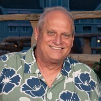 Casual outdoor portrait of Bill Lang, Premium Auditor in Travelers Business Insurance Operations. He has gray-white hair and displays a warm, friendly smile. He wears a blue and white tropical floral Hawaiian shirt, photographed in natural lighting.