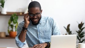 A man sitting at office desk in front of laptop hold mobile phone make pleasant business or informal call.