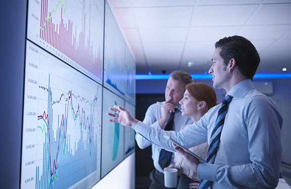 Three business people discuss graphs on screen in meeting room