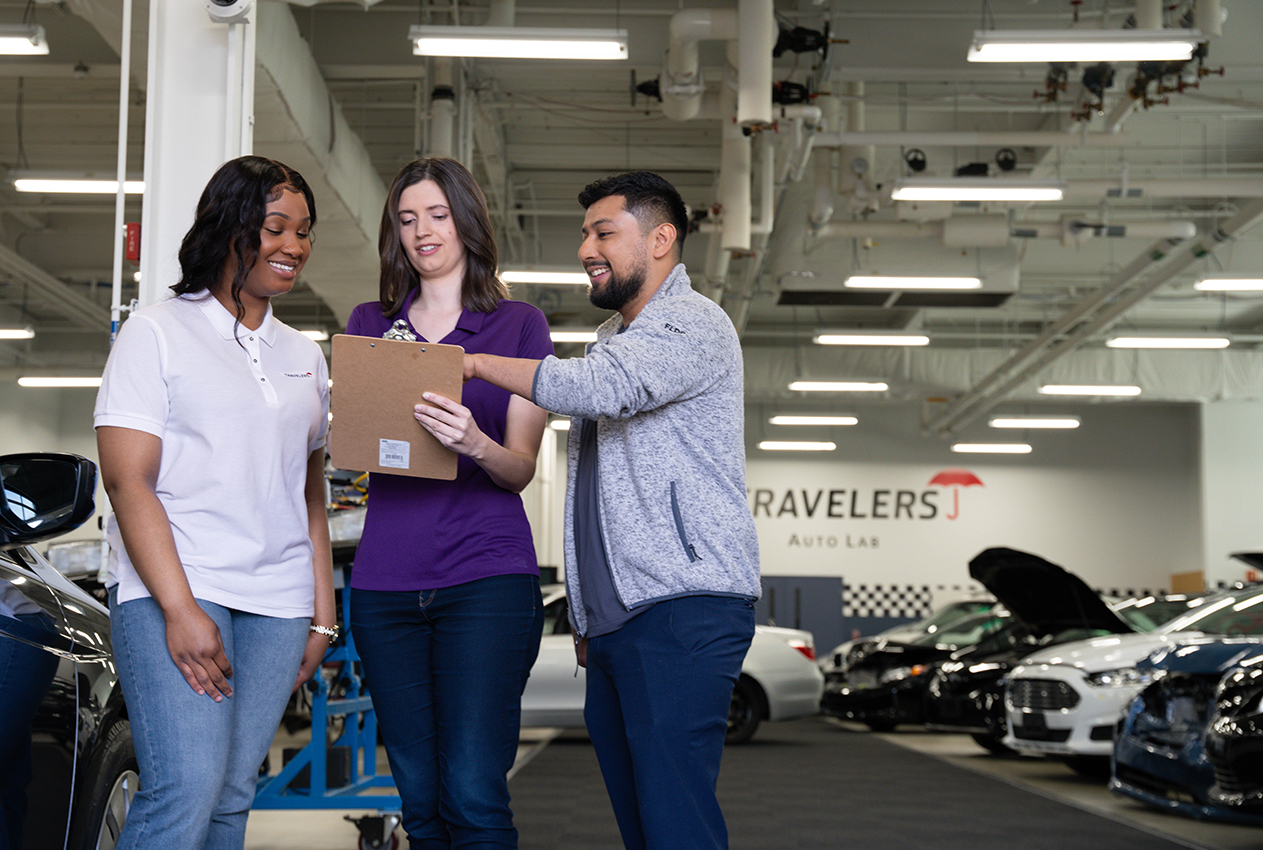 internshipTertiaryImage Three people stand together in an automotive service facility with an industrial ceiling and fluorescent lighting. They are reviewing a document or clipboard. On the left is a woman in a white polo shirt and jeans, in the middle is a woman in a purple top and jeans, and on the right is a man in a gray sweater and jeans. A 'TRAVELERS' sign is visible on the wall, and several cars are in the background.