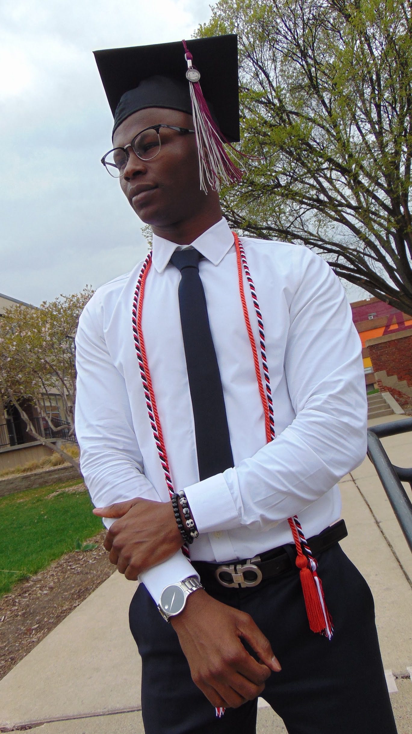 A person dressed in graduation attire stands outdoors in a campus setting. They are wearing a white dress shirt, black tie, and black pants, complemented by red and white honor cords around their neck. A graduation cap with a matching tassel adorns their head. The background features trees and a building, indicating a school or university environment. The individual accessorizes with a watch, bracelets, and a belt with a distinctive buckle.