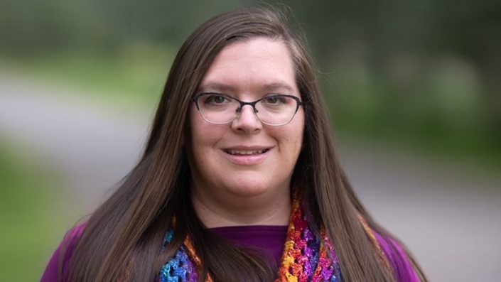 A woman with long, straight brown hair and glasses is smiling warmly. She is wearing a purple top and a vibrant, multicolored crocheted or knitted scarf with bright hues of blue, purple, orange, and yellow. The background is softly blurred, suggesting she is outdoors with greenery or trees behind her. The lighting is natural, giving the photo a professional quality.