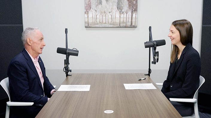 Two people sitting across from each other at a wooden conference table in a professional interview or podcast recording setup. On the left is an older man with gray hair wearing a dark suit, and on the right is a younger woman with long brown hair wearing a black blazer. Both have microphones positioned in front of them and papers on the table. The room has white walls with framed artwork and appears to be a modern office or studio space.