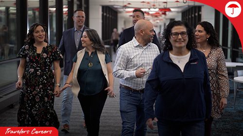 A group of seven professional colleagues walking together through a modern office hallway with glass walls and contemporary lighting. The group includes men and women of various ages dressed in business casual attire, appearing to be engaged in friendly conversation as they move through the workspace. The scene conveys a collaborative, inclusive workplace environment."