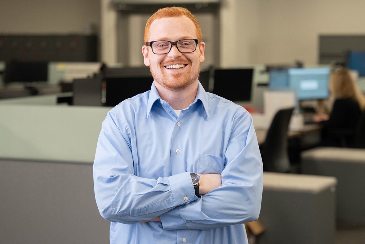 ETP Photo_resized_2 A professional headshot of a smiling man with red/ginger hair and black-rimmed glasses, wearing a light blue button-down shirt. He is standing with his arms crossed, exuding confidence. A dark-colored watch is visible on his wrist. The background features a modern open office space with cubicles, computer monitors, and other employees working at their desks.