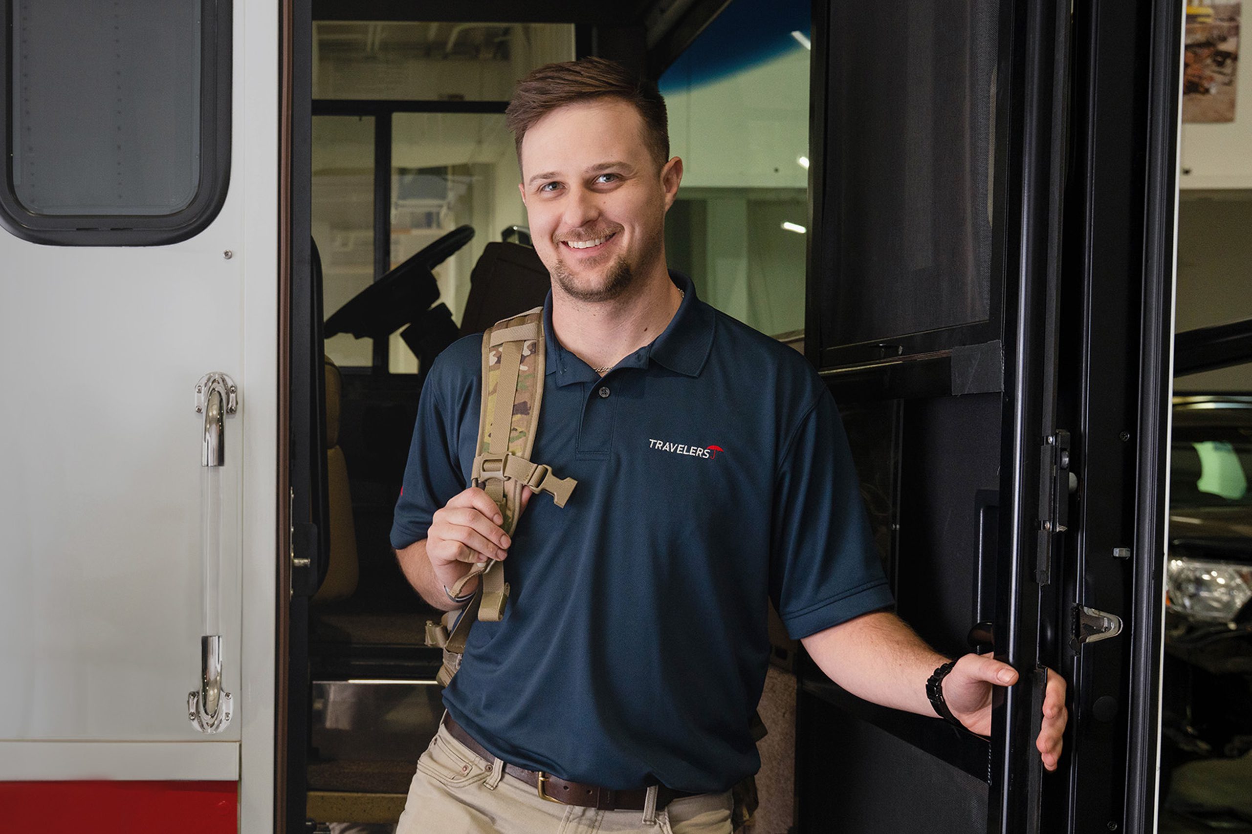 Interested-in-Learning-More-Military A smiling man in a navy blue polo shirt stands in the doorway of what appears to be an RV or motorhome. He's holding a backpack with a tan/desert camouflage pattern and has his other hand on the door frame. The interior of the vehicle is visible behind him, showing a modern, well-lit living space.
