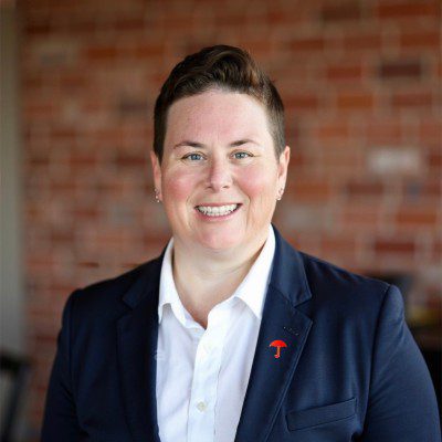 Professional headshot of Tiffany Bilbrey, Personal Insurance Operations Customer Service Representative at Travelers, smiling warmly in a navy blazer and white shirt with a red lapel pin, photographed against an exposed brick wall background.