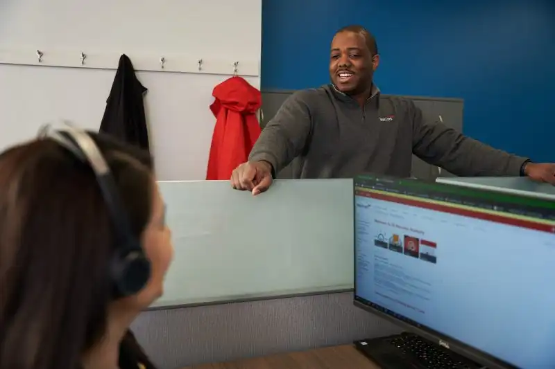 A smiling man in a gray sweater stands behind a white cubicle partition in an office setting, appearing to have a friendly conversation with a colleague who is seated at a desk with a laptop computer. The office has a blue accent wall with coat hooks holding a black jacket and red hoodie. 