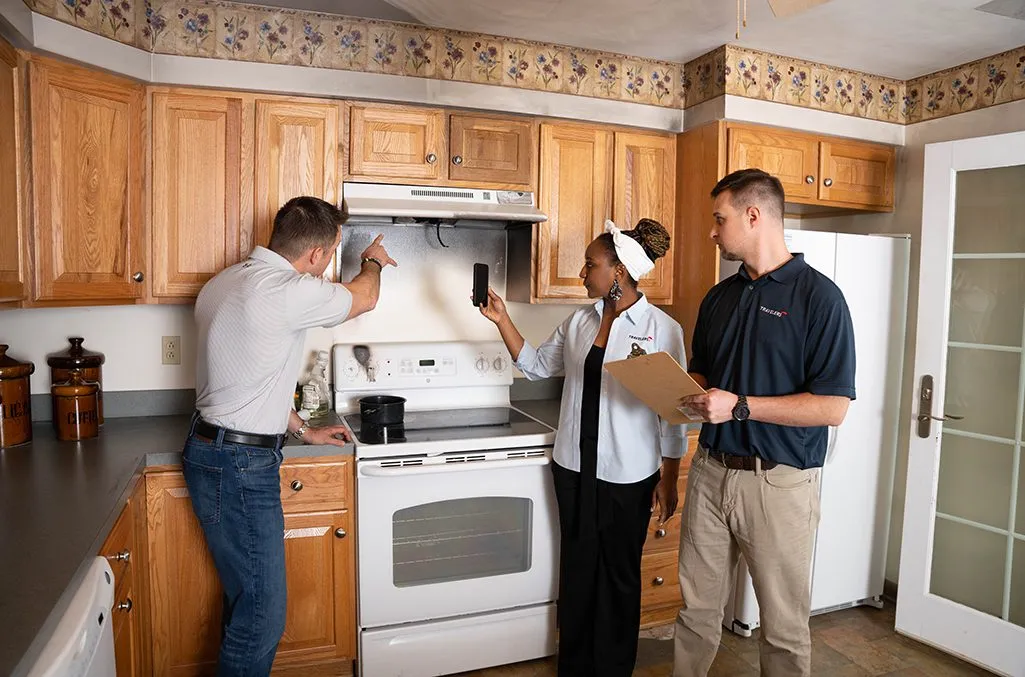 Claim-secondary Three people conducting a home inspection in a kitchen with wood cabinets. One inspector on the left examines the range hood while holding a tool, a woman in the center points upward wearing a white shirt and cap, and a male inspector on the right holds a clipboard. The kitchen features honey-colored wood cabinets, white appliances, dark countertops, and decorative wallpaper border.