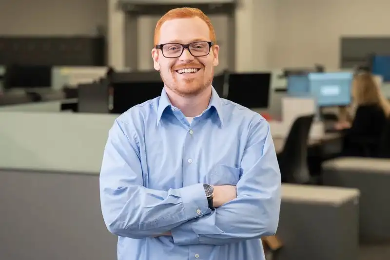 A professional headshot of a smiling man with red/ginger hair and black-rimmed glasses, wearing a light blue button-down shirt. He is standing with his arms crossed, exuding confidence. A dark-colored watch is visible on his wrist. The background features a modern open office space with cubicles, computer monitors, and other employees working at their desks.