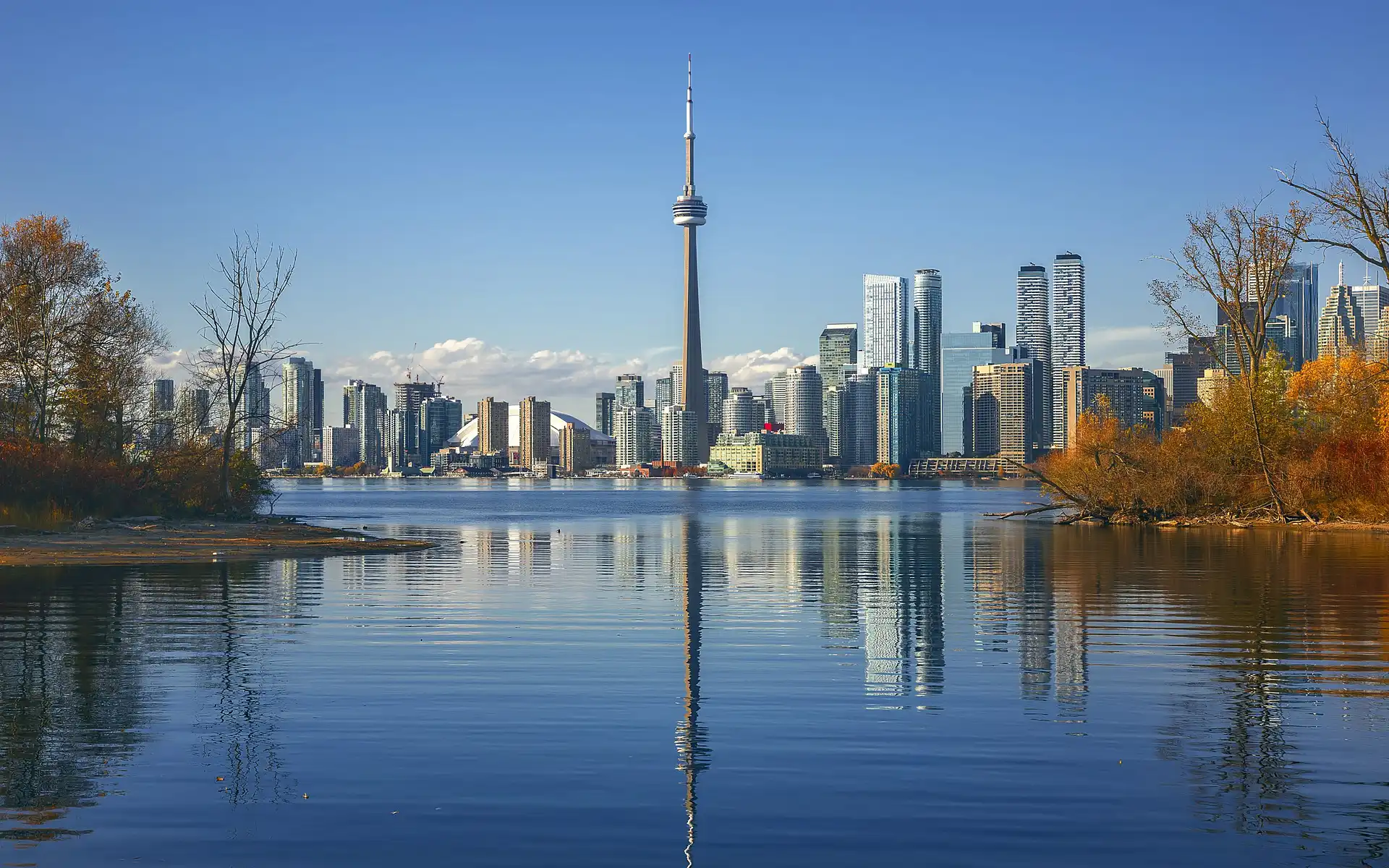 Photograph of Toronto's skyline taken from across the water, featuring the iconic CN Tower prominently in the center, surrounded by modern skyscrapers. The foreground includes calm water reflecting the skyline and autumn-colored trees with golden and orange foliage. The sky is clear blue with some white clouds, and the lighting suggests it was taken during golden hour, giving the scene a warm glow.