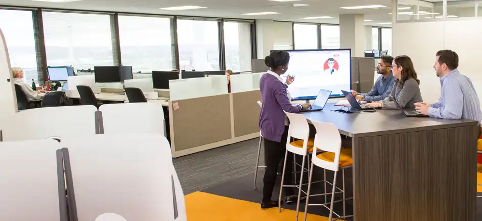 Three people sit and one stands at a tall table in an office, looking at a screen together